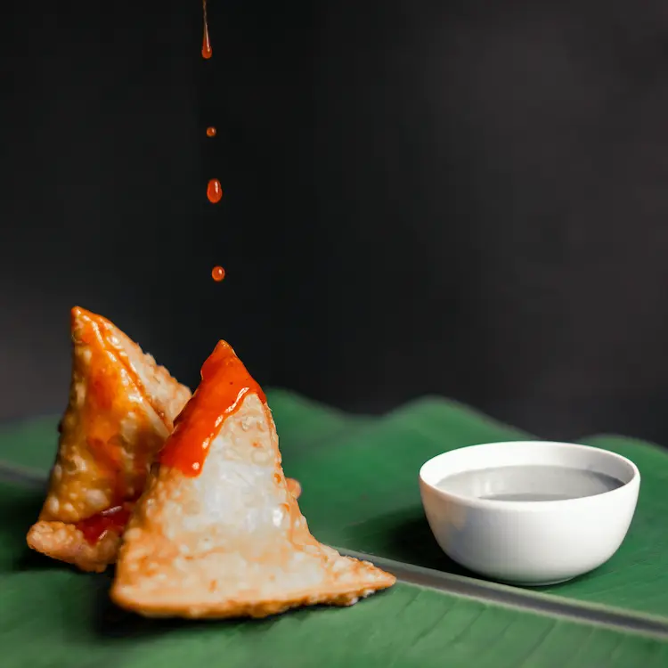 Close-up of two crispy samosas being drizzled with tangy red sauce, placed on a green leaf with a side of dipping sauce in a white bowl.