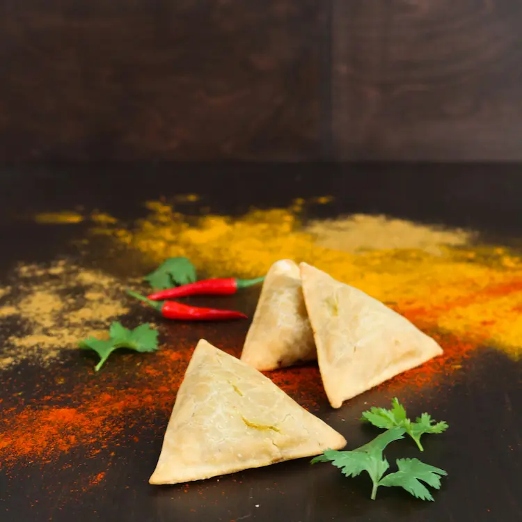 Two freshly baked samosas displayed on a dark surface with vibrant chili peppers, coriander, and colorful spice powders scattered around.