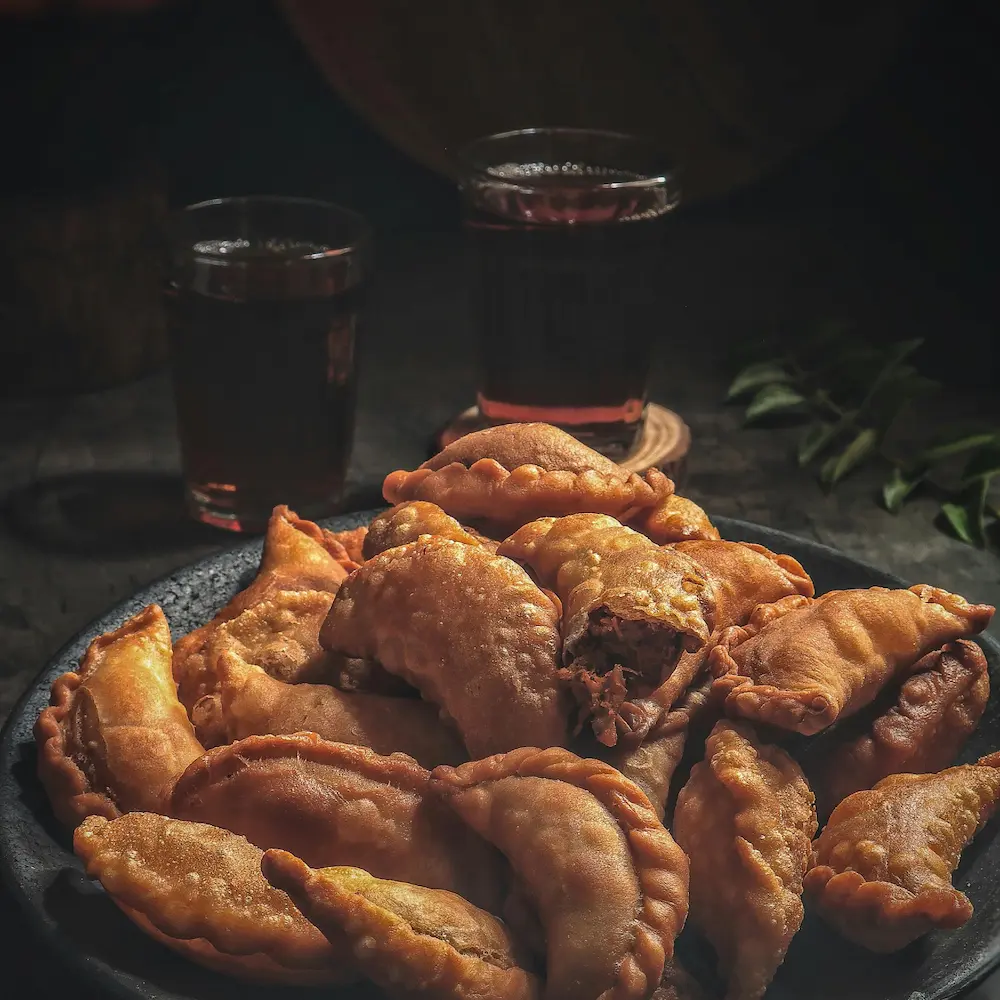 A rustic black plate filled with golden, crispy fried samosas, surrounded by two glasses of tea.