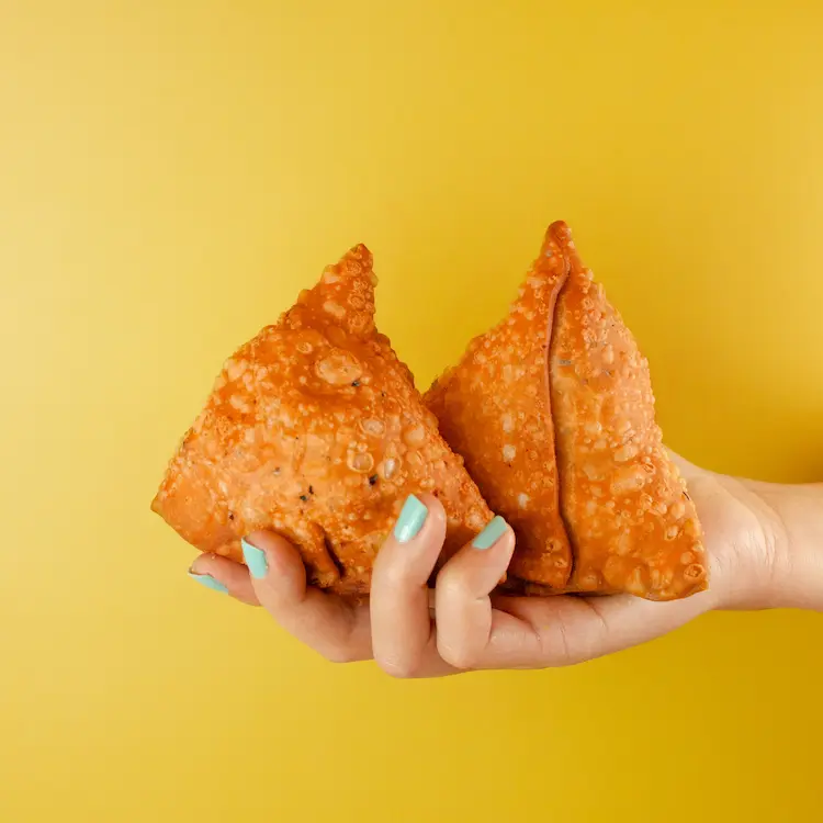 A hand holding two golden, crispy samosas against a bright yellow background, showcasing their perfect texture.