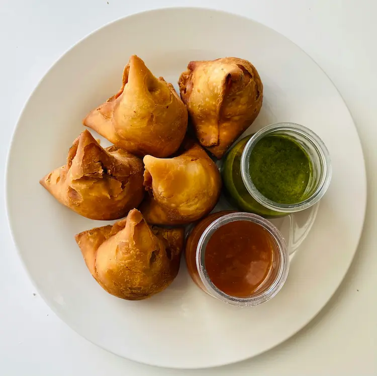 A white plate with 5 golden, crispy samosas accompanied by jars of green mint chutney and brown tamarind sauce.